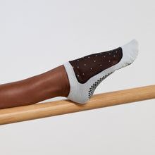 Cargar imagen en el visor de la galería, Close-up of a foot wearing white grip socks with a sheer black mesh panel and small dotted details, resting on a wooden barre.
